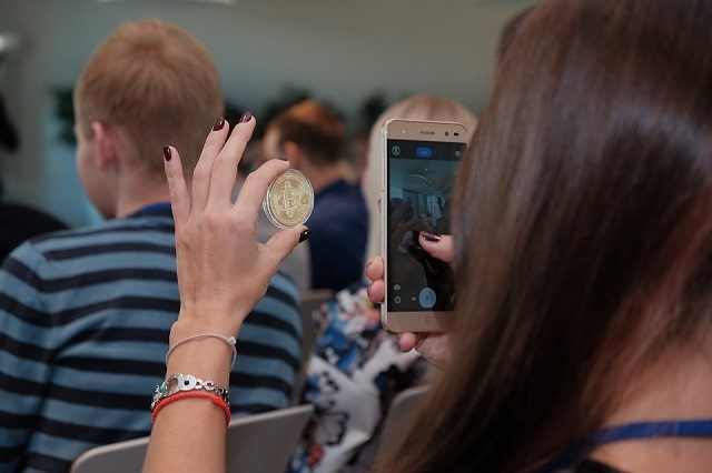 kobieta robi zdjęcie trzymanemu bitcoin tło stanowią inne osoby sala wykładowa